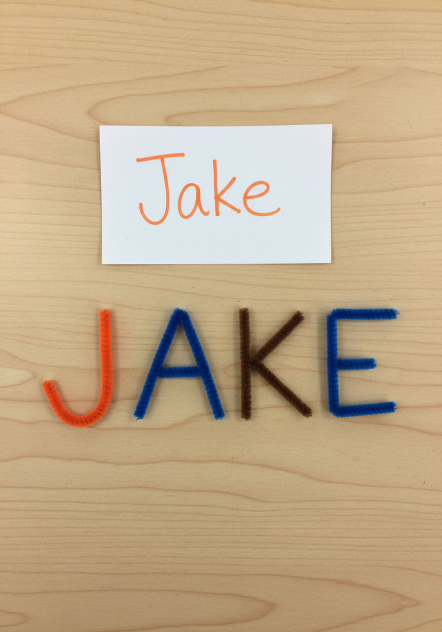 Completed name Jake spelled out using colorful pipe cleaners placed below index card with written name for build a name STEM activity