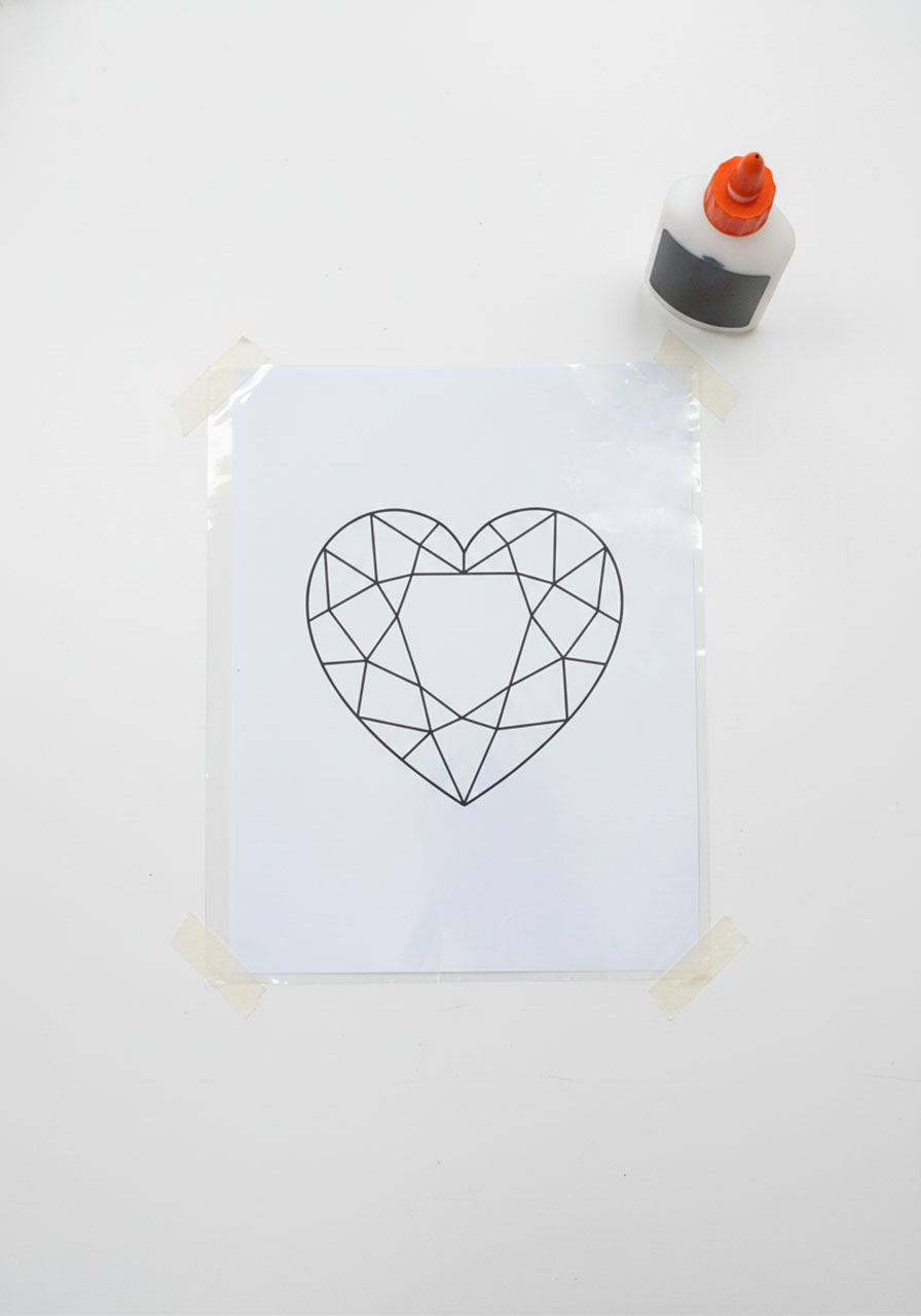Geometric heart template placed under clear plastic sheet, taped down and ready for black glue tracing to make stained glass suncatcher