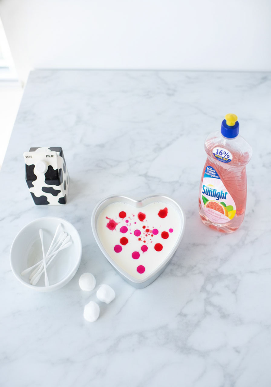 Valentine's Day magic milk heart science experiment setup with red and pink food coloring drops in heart-shaped pan with dish soap and cotton swabs for kids
