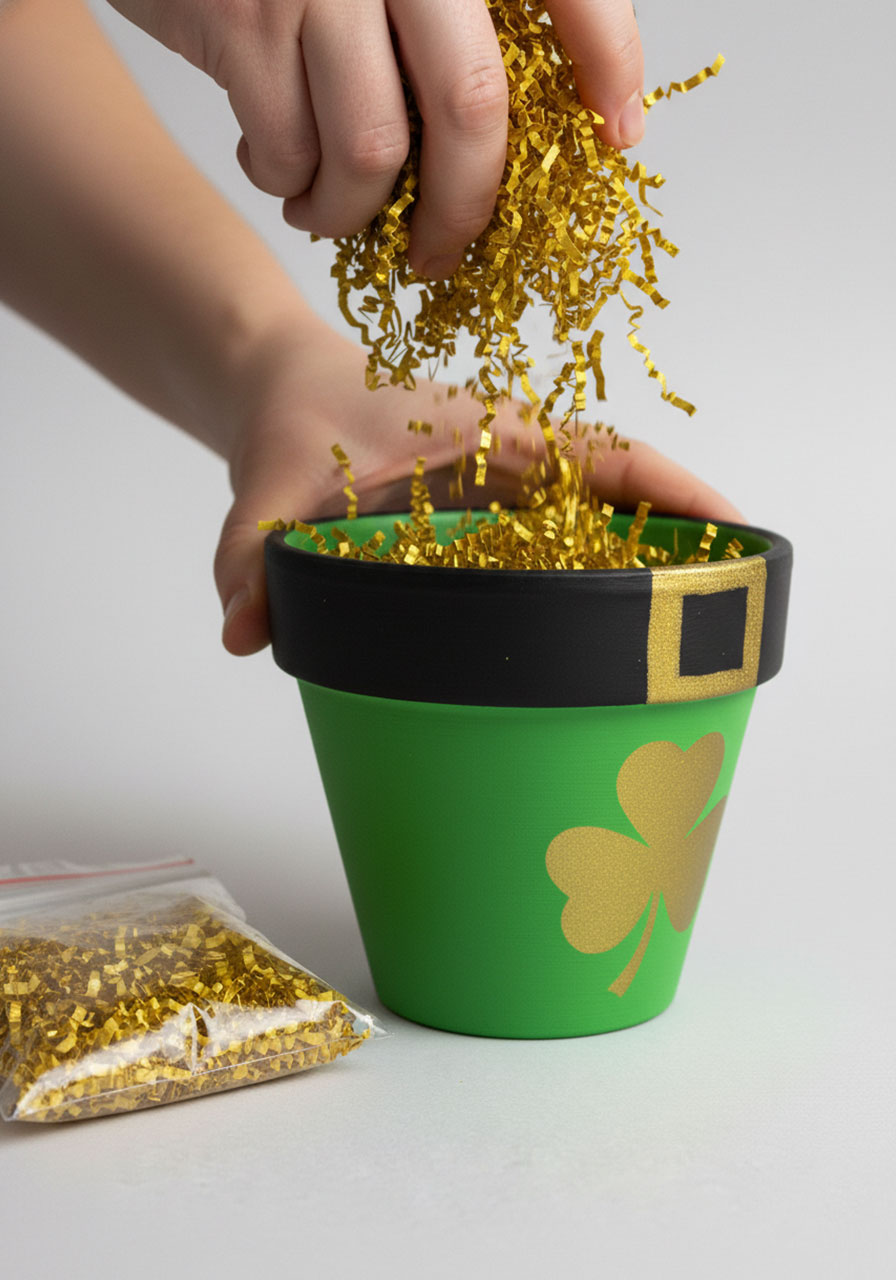 Hands adding gold crinkle paper filler to leprechaun pot before adding candy for St Patrick's Day gift