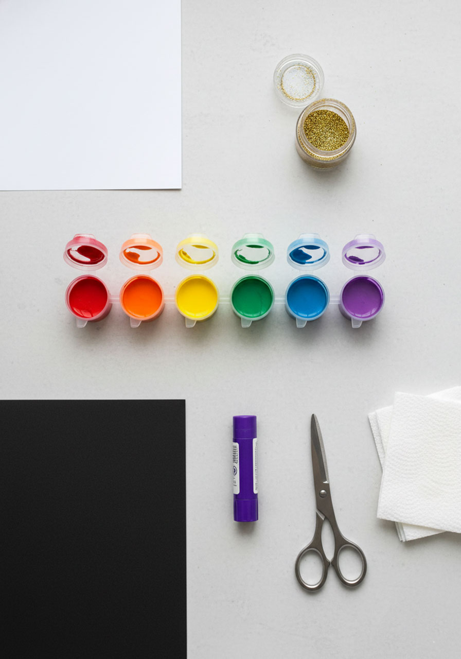 Flat lay of supplies needed for fingerprint rainbow craft including paints, paper, glitter, glue stick, and scissors