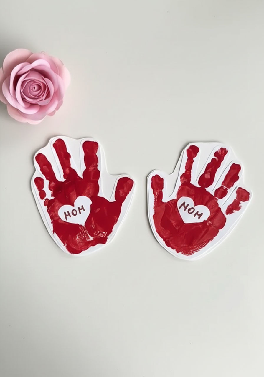 Finished DIY Mother's Day handprint card with red painted handprints and white heart centers saying MOM, shown closed before the accordion message is revealed