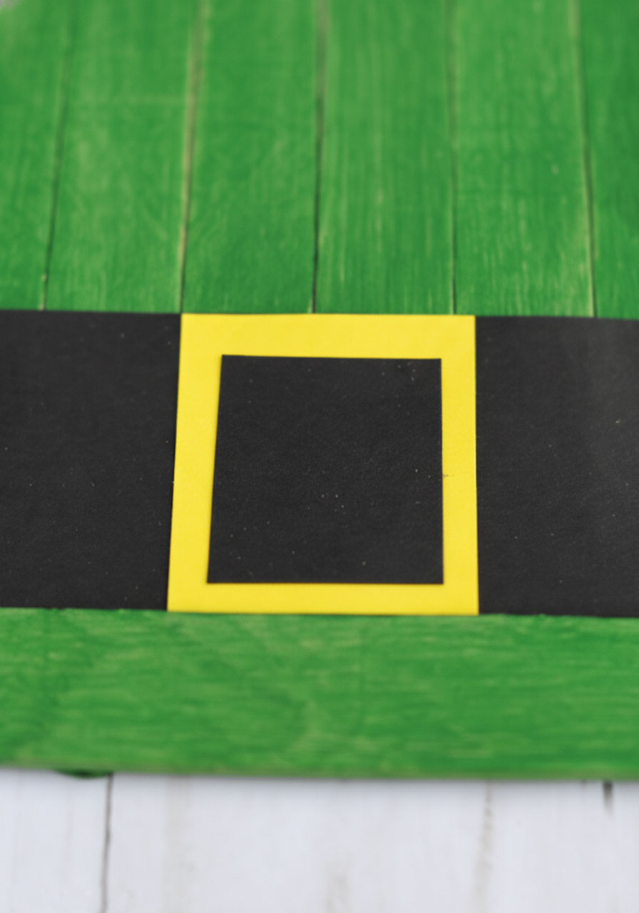 Close-up of black paper belt with yellow and black layered square buckle on green craft stick leprechaun hat