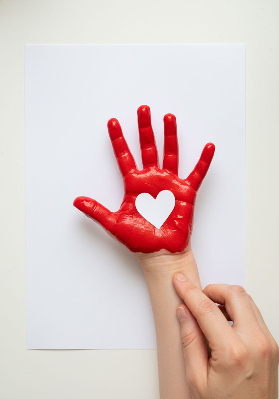 Child's painted red hand being pressed onto white cardstock with a small paper heart placed in the palm center to create a heart negative space in the handprint