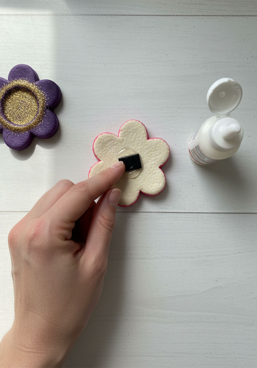 Adult pressing a small black craft magnet onto the back of a finished pink salt dough flower magnet with craft glue