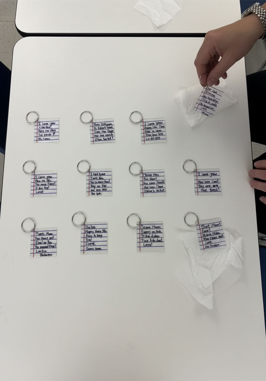 Twelve completed DIY shrink plastic notebook paper keychains spread on classroom table made by students for Mother's Day