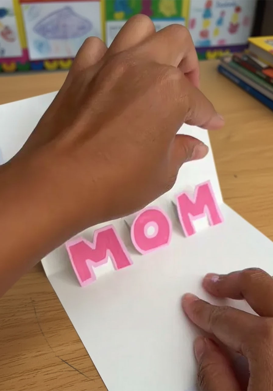 Adult hands gluing pink block letter M onto a pop-up tab inside an open handmade birthday card with completed O letter already standing on center tab step4b