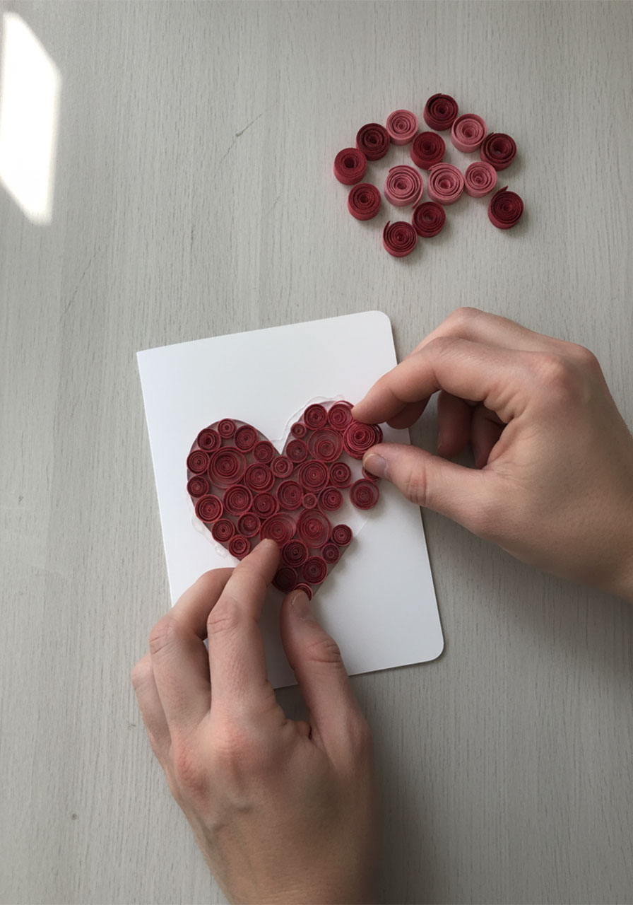 Hands placing rolled pink and red paper quilling spirals into heart-shaped window of handmade Mother's Day card