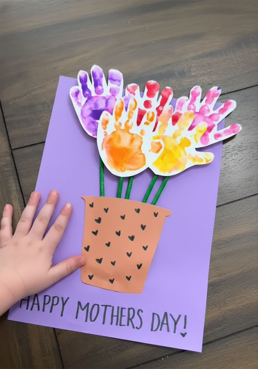 Finished DIY handprint flower bouquet Mother's Day card with colorful toddler handprints on green stems in paper pot on purple background with happy mothers day message
