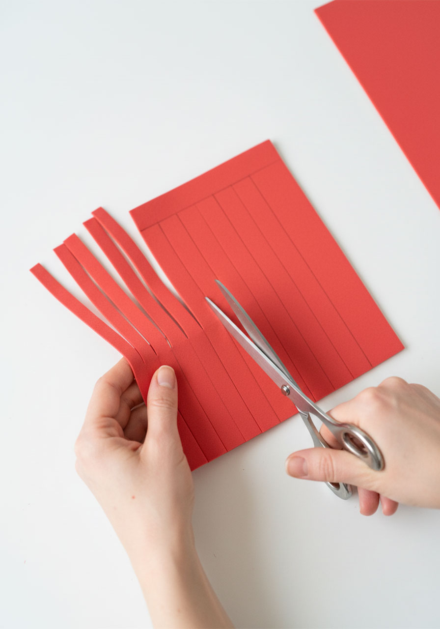 Adult hands cutting vertical fringe strips into a red foam sheet with scissors to make DIY car wash curtain for tissue box craft