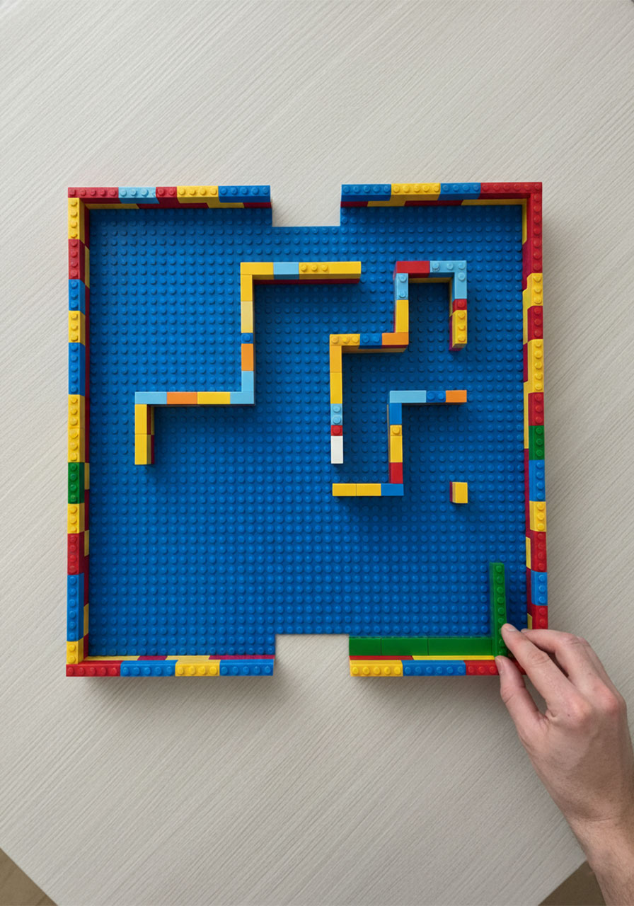 Adult hand placing green LEGO bricks inside a baseplate border to build the interior walls of a marble maze