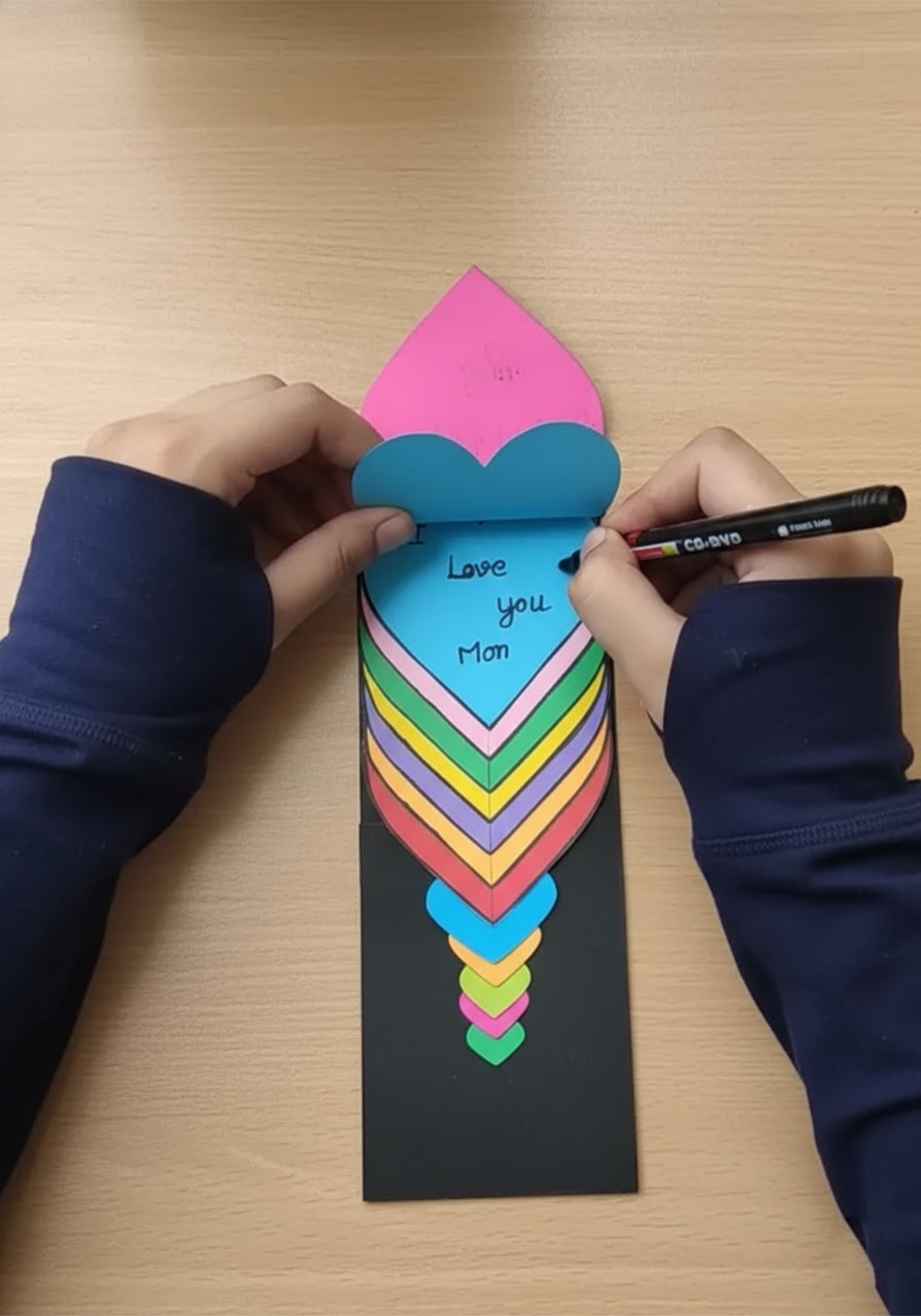 Child writing "I Love You Mom" on a paper heart for a handmade Mother's Day heart waterfall card