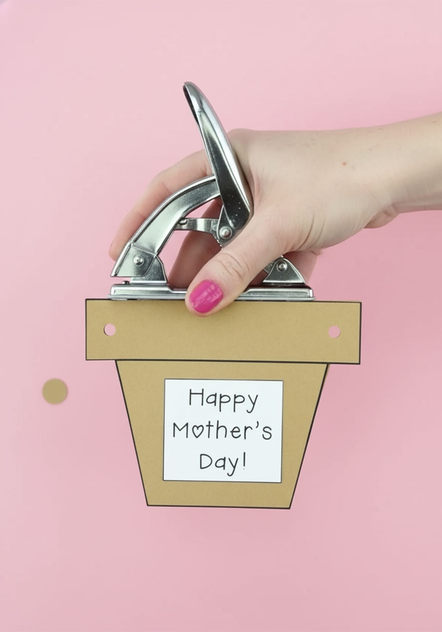 Using a hole punch to make two ribbon holes in the top rim of a brown kraft cardstock flower pot Mother's Day card