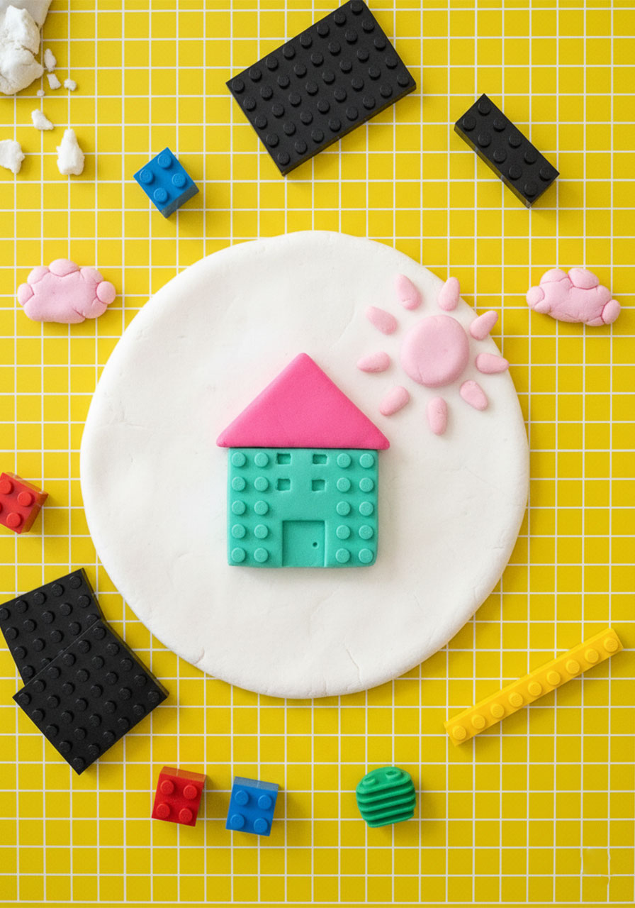 Overhead flat-lay of white playdough canvas with colorful playdough house and sun collage scene created using LEGO brick stamps, surrounded by LEGO pieces on yellow craft mat