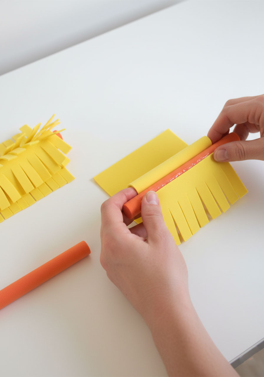 Adult hands rolling yellow fringed foam sheet around an orange foam stick to create a spinning brush roller for DIY tissue box toy car wash