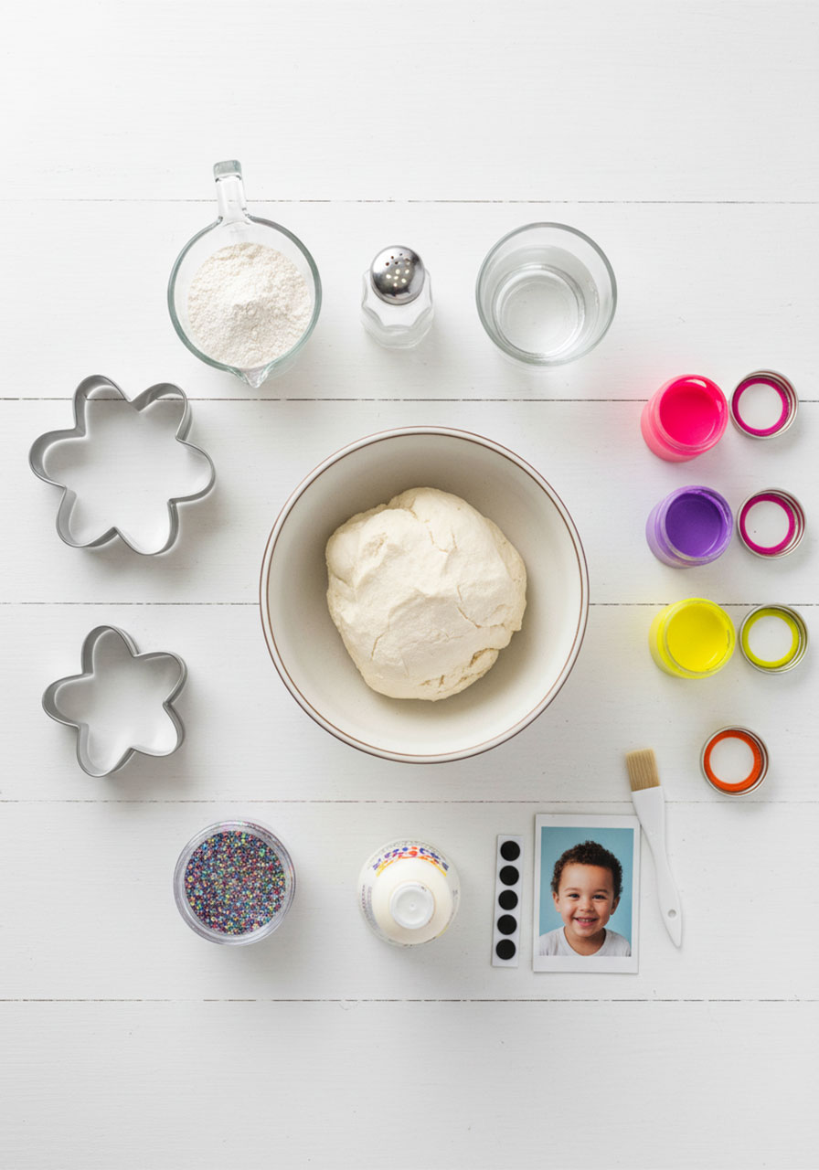 Flat lay of salt dough flower magnet supplies including cookie cutters, paint, glitter, Mod Podge, magnets, and a child's photo on a white surface