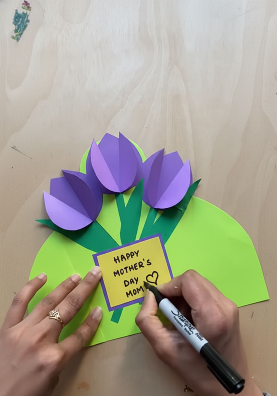 Writing "Happy Mother's Day Mom" with a black marker on a small yellow message tag card for a handmade paper tulip card