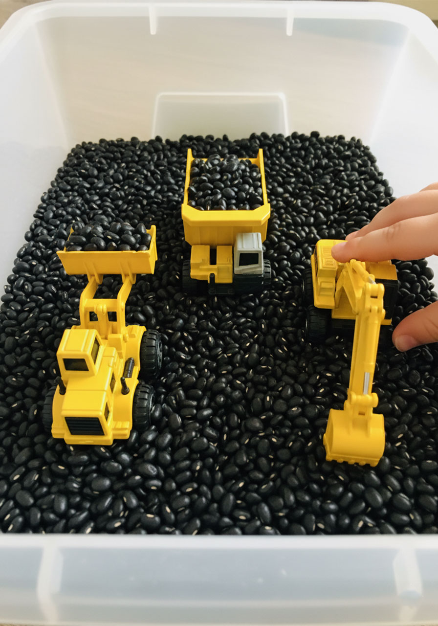 Small yellow toy construction trucks including dump truck and excavator surrounded by black beans in a sensory bin