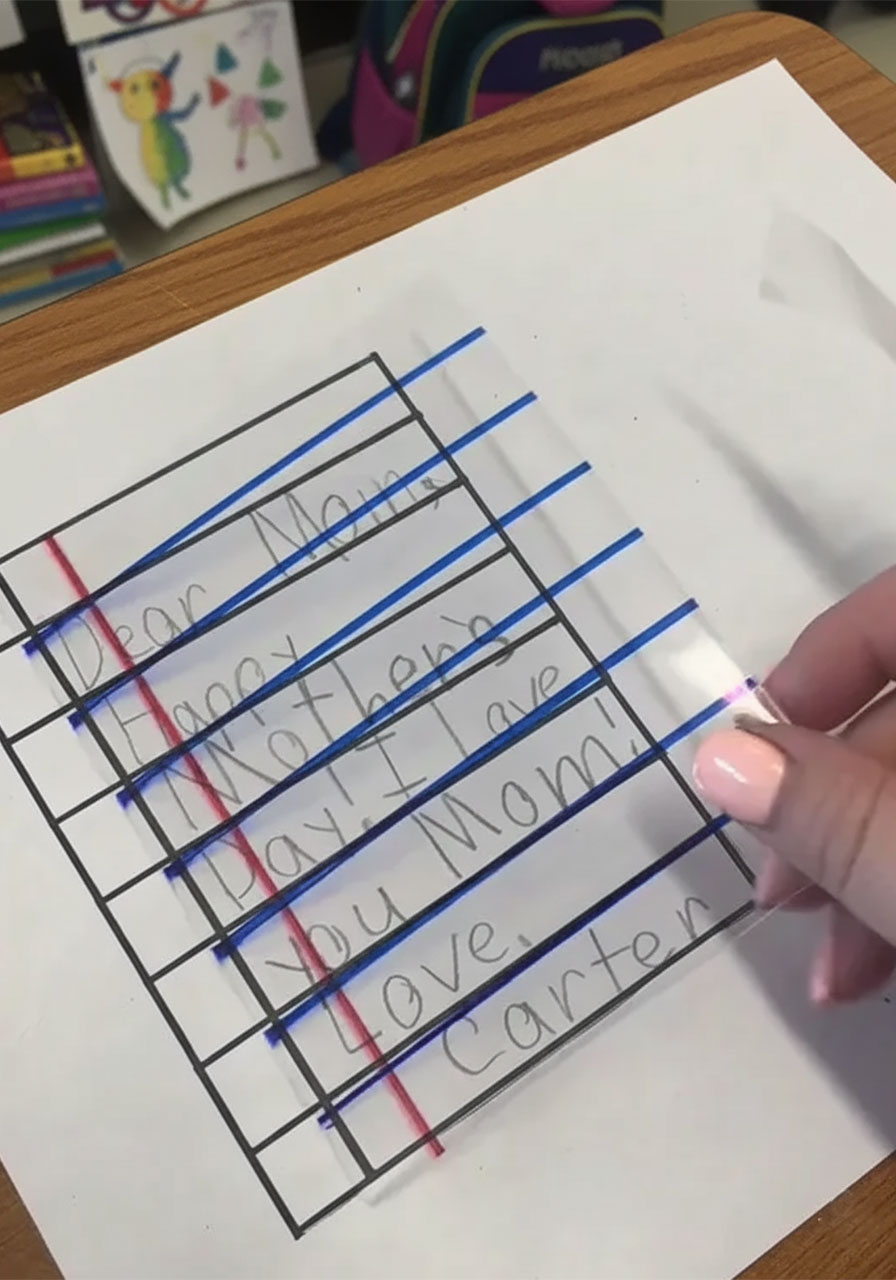 Adult tracing child's handwritten Mother's Day message onto clear shrink plastic sheet with black Sharpie over notebook paper template step3a