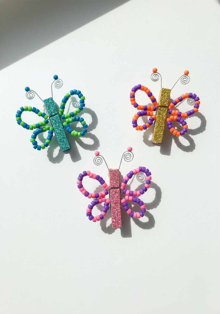 Three completed beaded pipe cleaner butterflies in pink, blue, and purple pony beads with glitter clothespin bodies on white surface