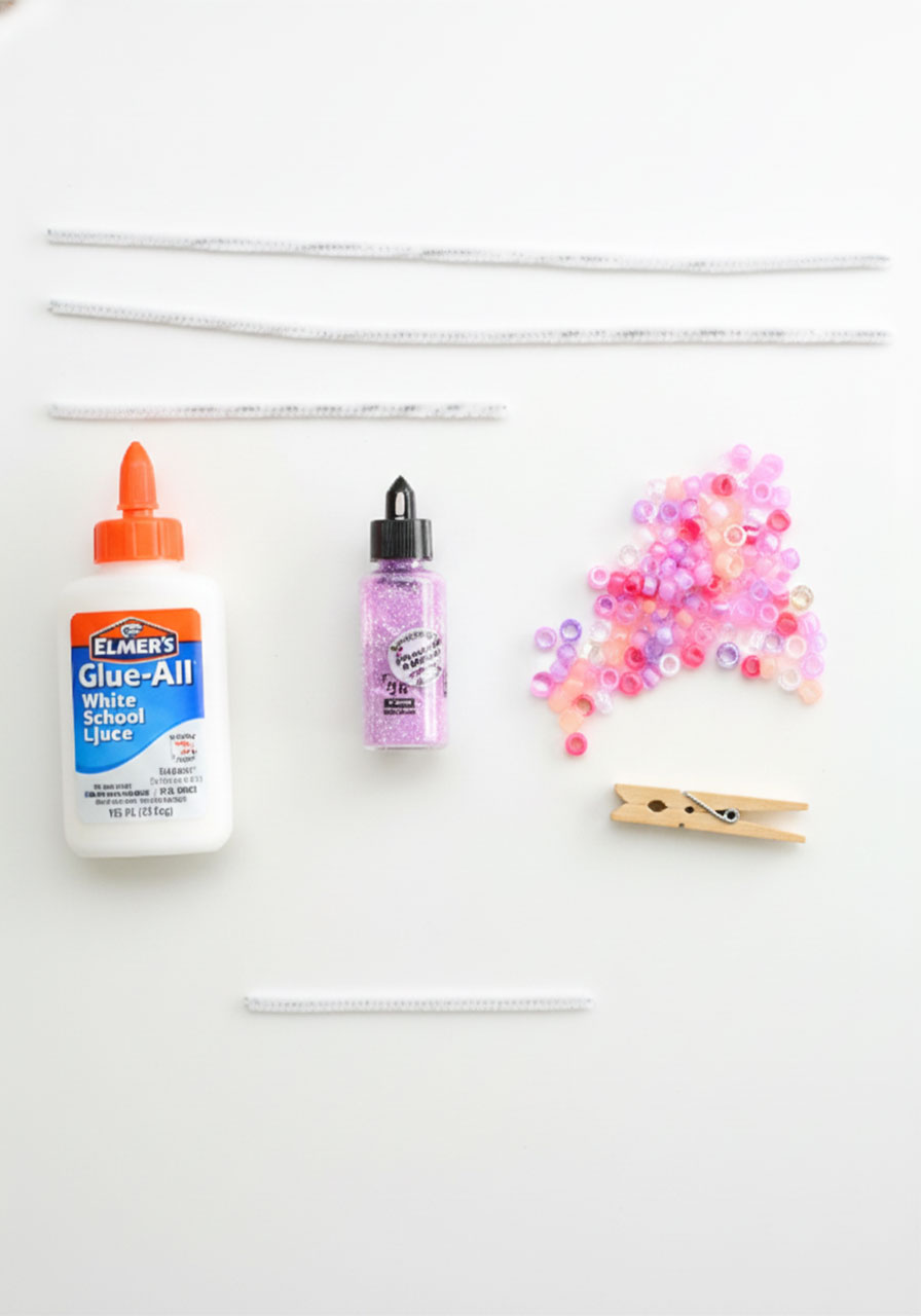 All supplies needed for beaded pipe cleaner butterflies including pipe cleaners, pony beads, clothespin, white glue, and glitter laid flat on white surface