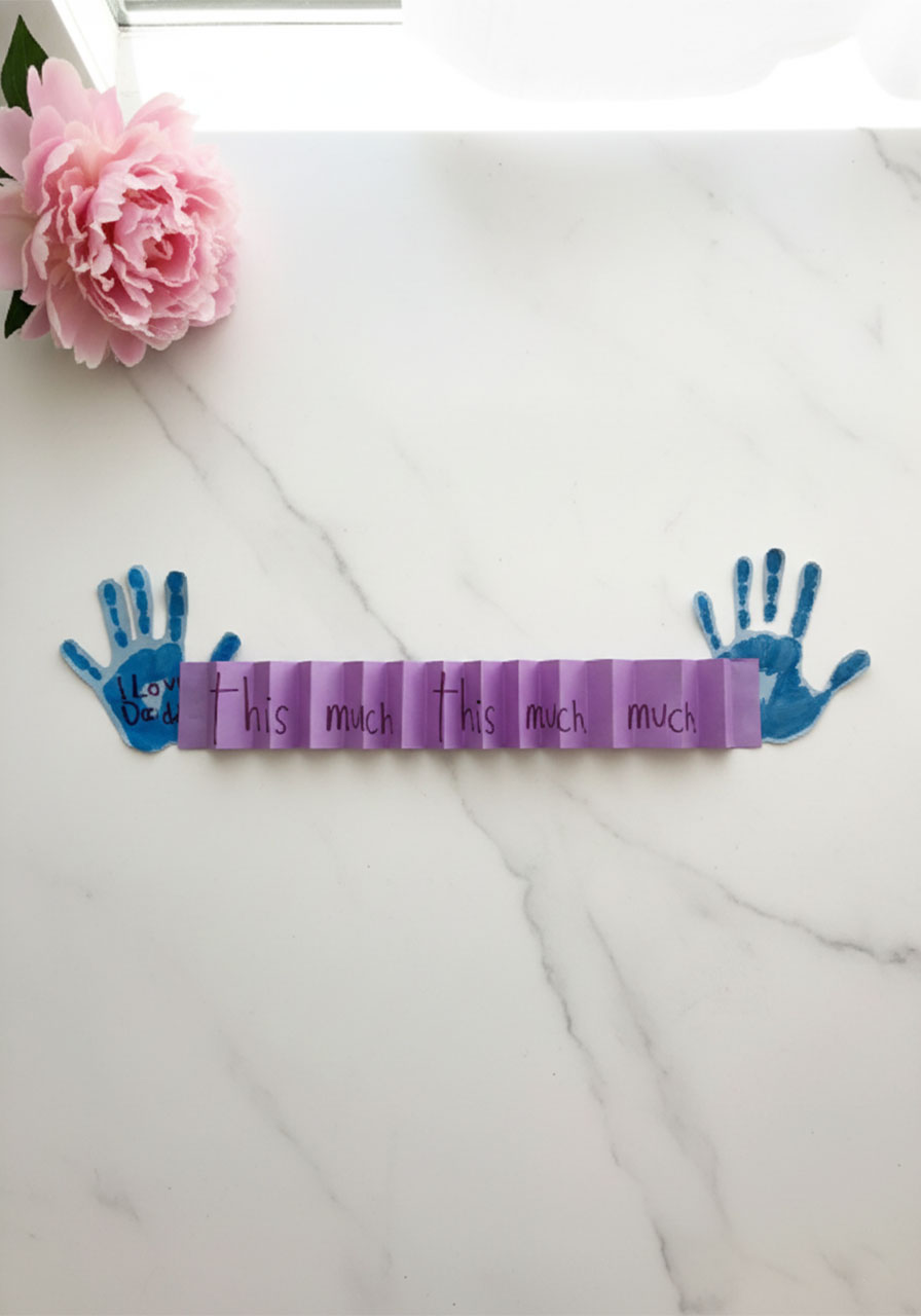 DIY I Love You This Much Father's Day handprint card displayed flat showing blue handprints and purple accordion strip reading this much