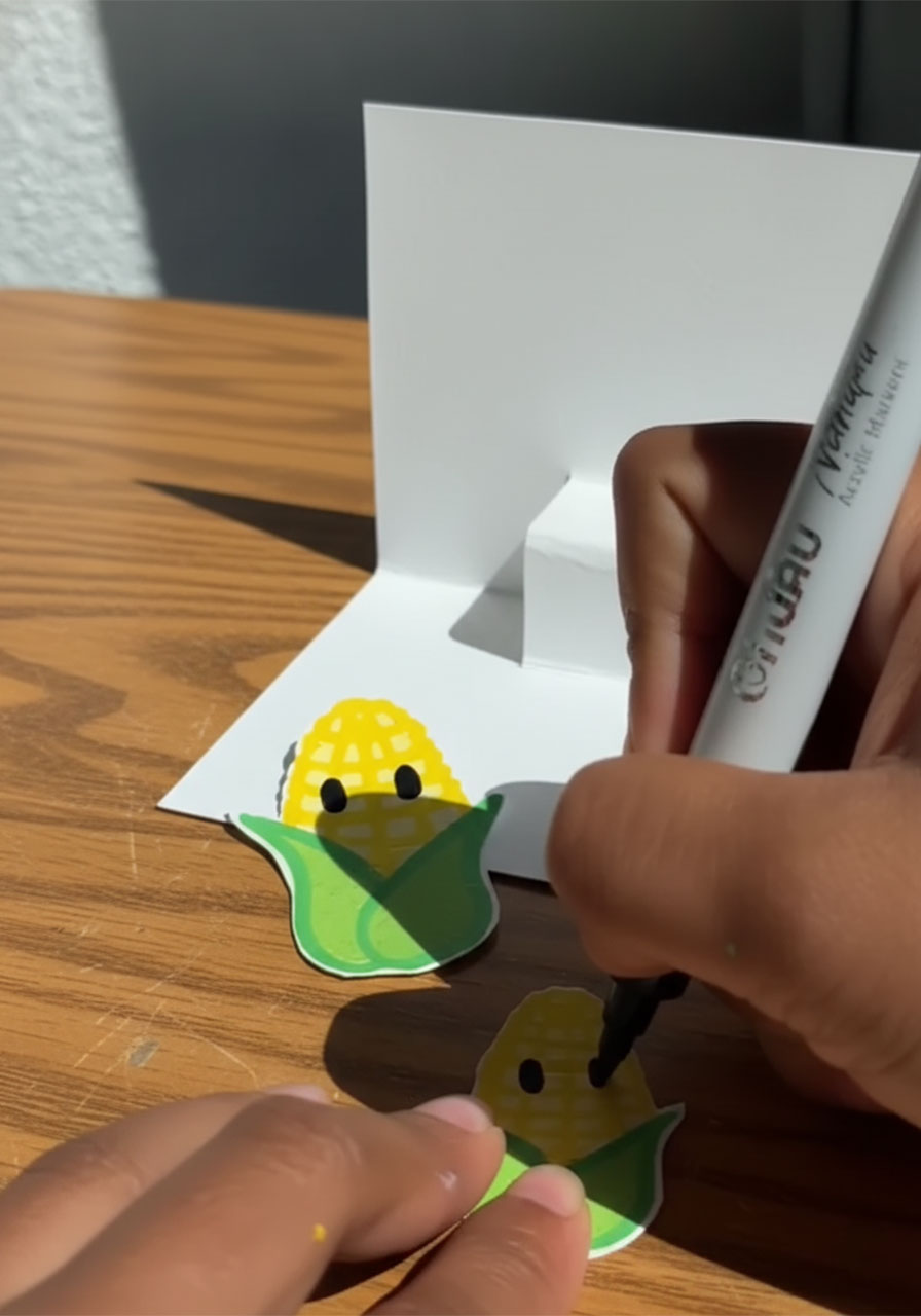 Child's hands drawing cute cartoon corn cob characters on white paper for a Father's Day pop-up card step2c