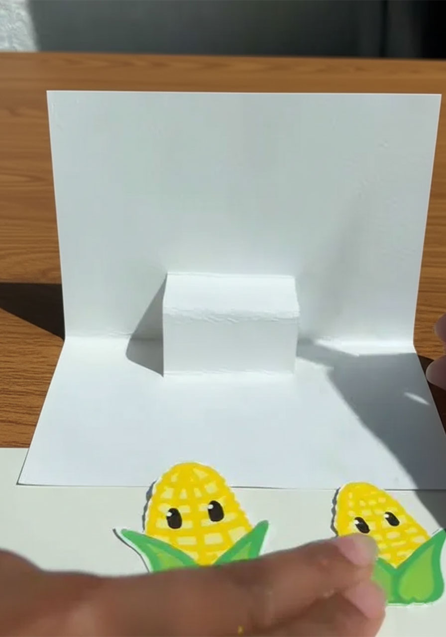Child's hands drawing cute cartoon corn cob characters on white paper for a Father's Day pop-up card step2d