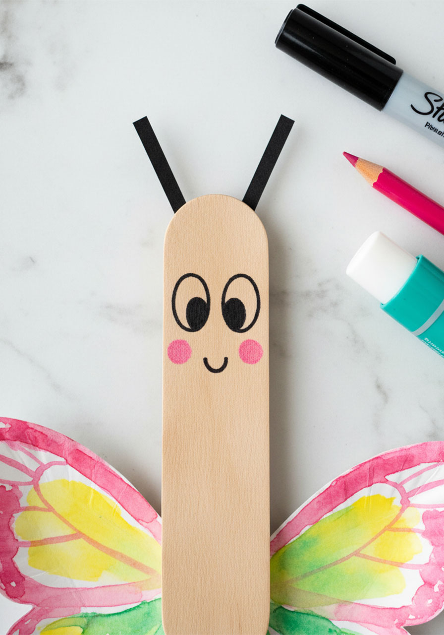 Close-up of a popsicle stick with a hand-drawn smiling butterfly face and black cardstock antennae glued to the top