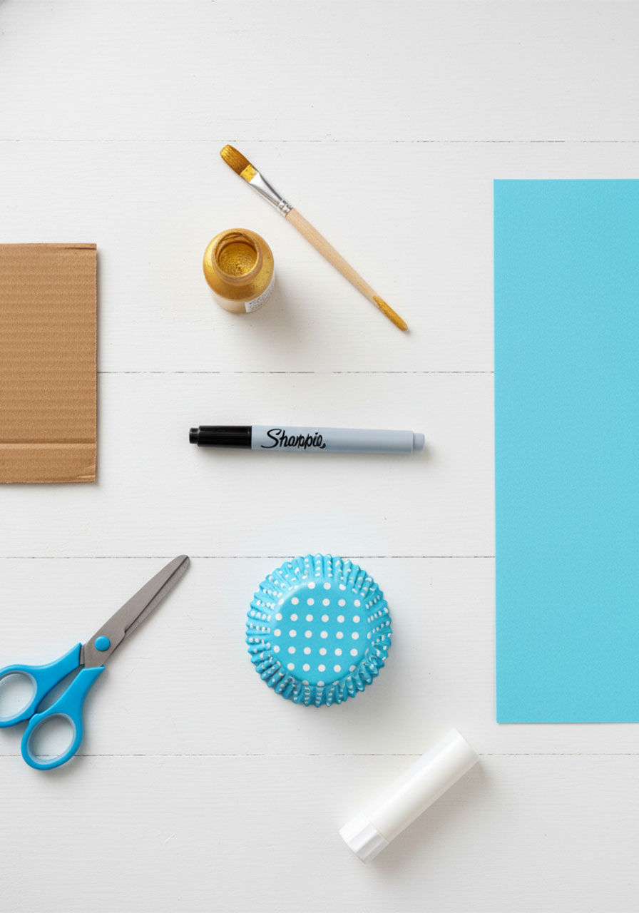 Flat-lay of all supplies needed to make a DIY Father's Day award ribbon including cardboard, gold paint, black marker, cupcake liner, blue construction paper, scissors, and glue