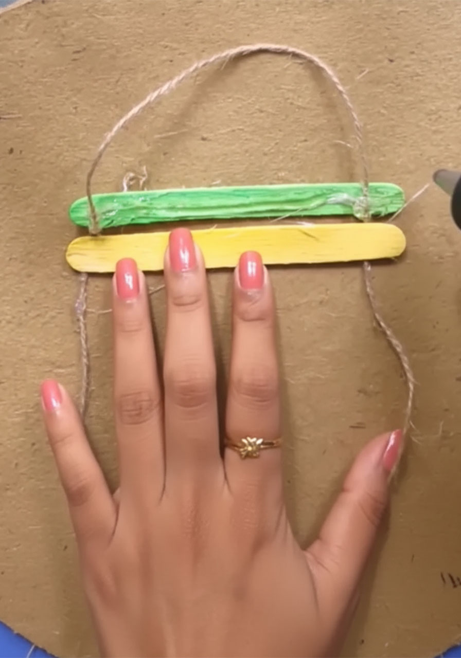 Adult hand hot gluing first green popsicle stick across jute twine to start Father's Day wall hanging craft project step2c
