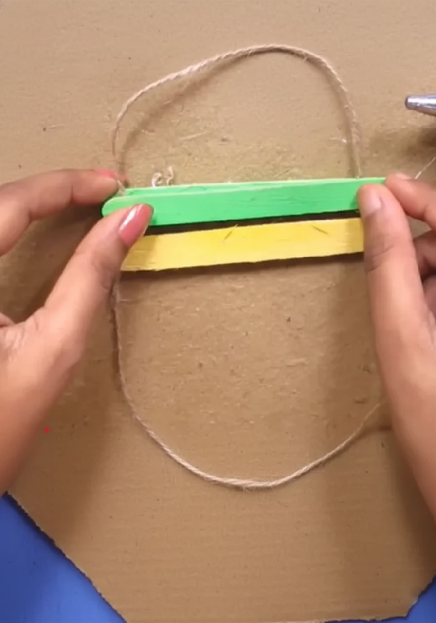 Adult hand hot gluing first green popsicle stick across jute twine to start Father's Day wall hanging craft project step2d