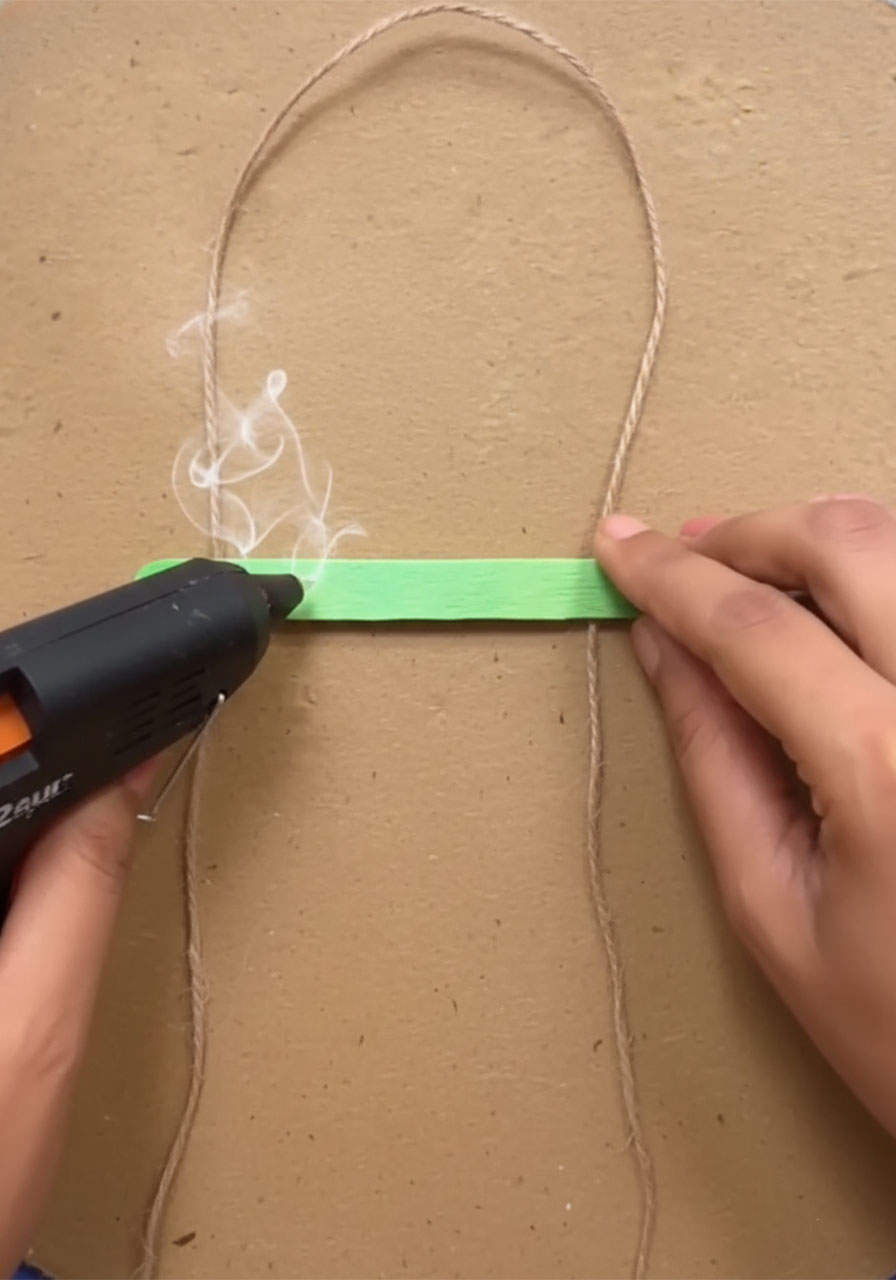 Adult hand hot gluing first green popsicle stick across jute twine to start Father's Day wall hanging craft project