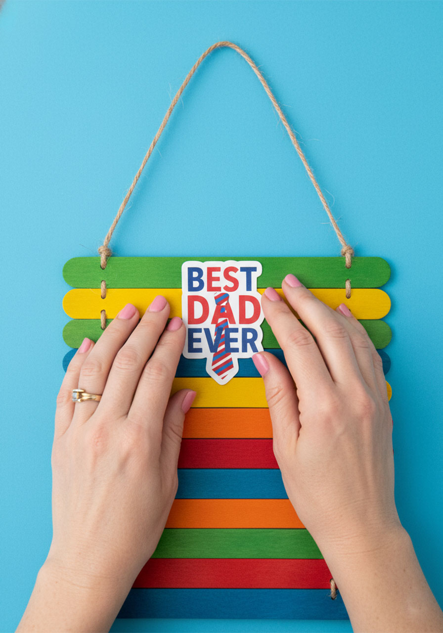 Pressing a Best Dad Ever sticker onto the top popsicle stick to decorate the Father's Day wall hanging craft