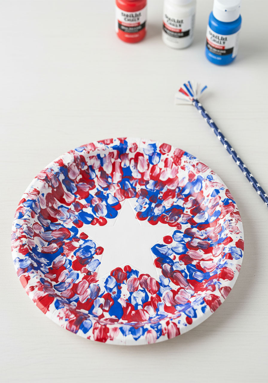 Single finished straw stamped paper plate 4th of July craft with white star cutout surrounded by red white and blue firework burst paint stamps