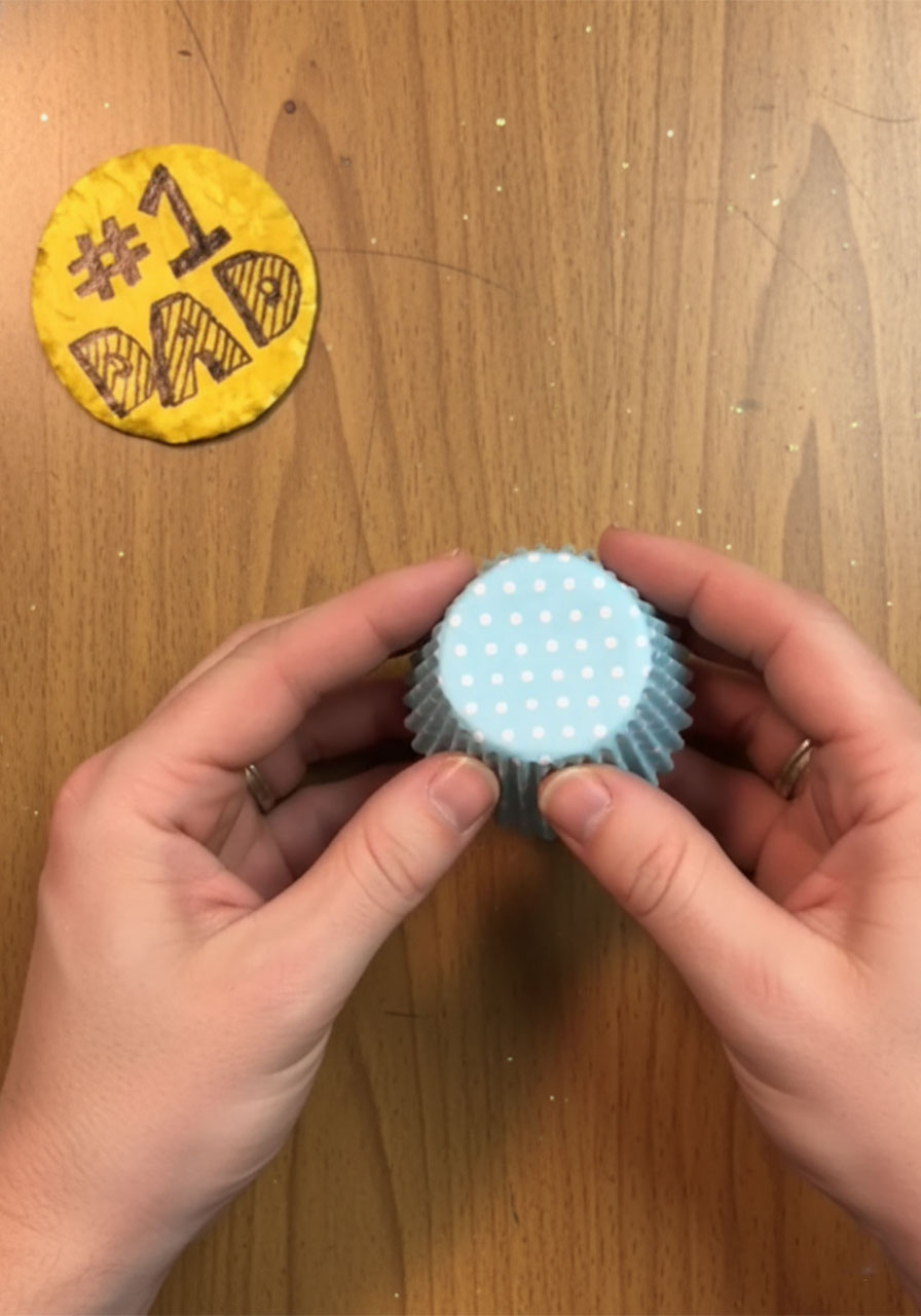 Child's hands flattening a blue polka dot cupcake liner to create a rosette frame for a DIY Father's Day award ribbon, Step 4a