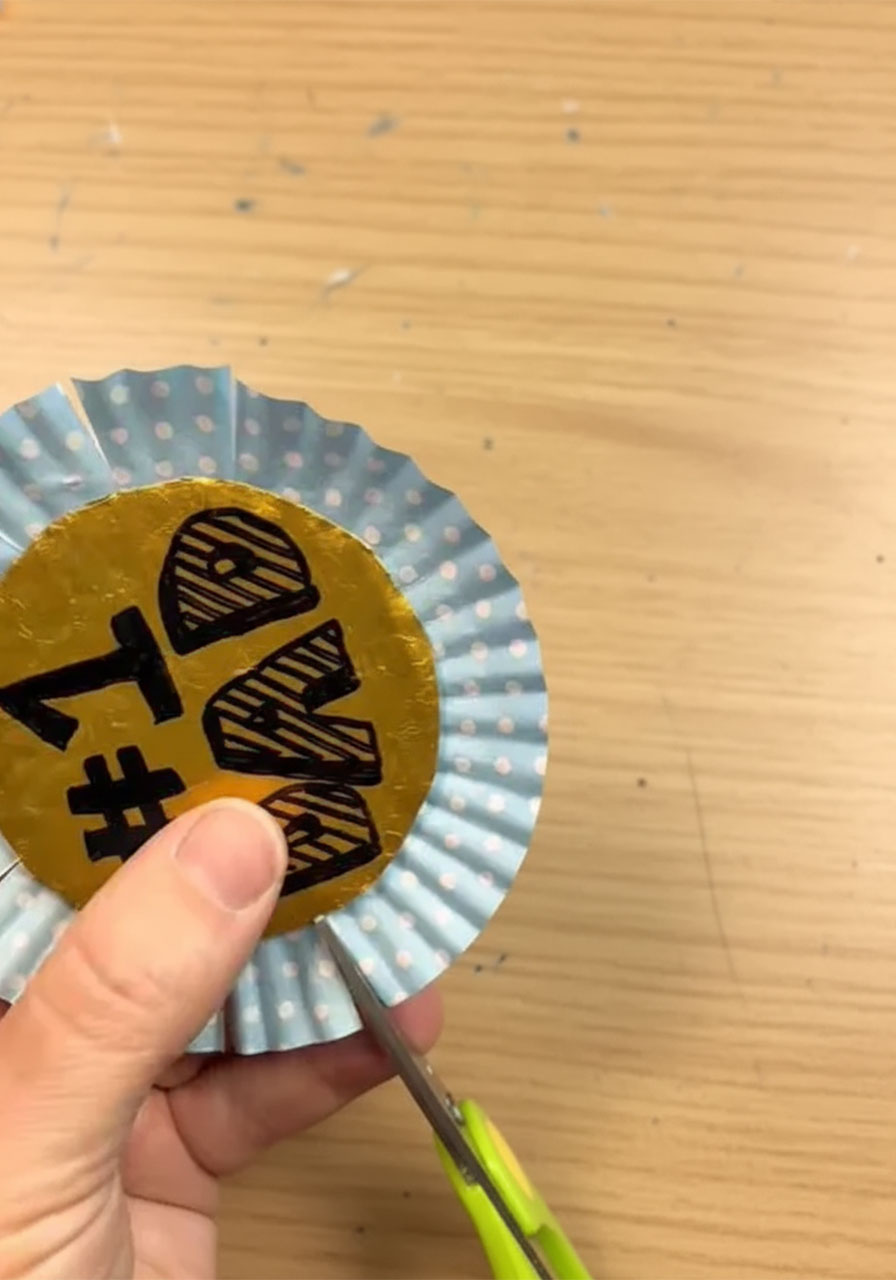 Child's hands flattening a blue polka dot cupcake liner to create a rosette frame for a DIY Father's Day award ribbon, Step 4b