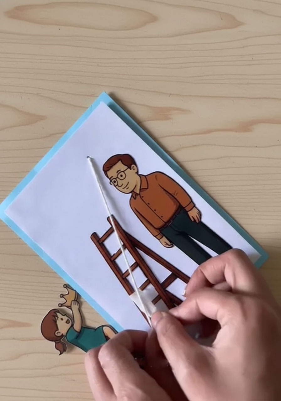 Adult hands gluing paper child figure onto pull-string of DIY Father's Day card with tiny crown ready to attach step6a