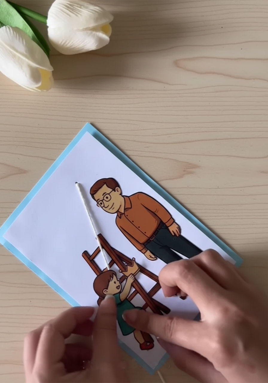 Adult hands gluing paper child figure onto pull-string of DIY Father's Day card with tiny crown ready to attach step6b