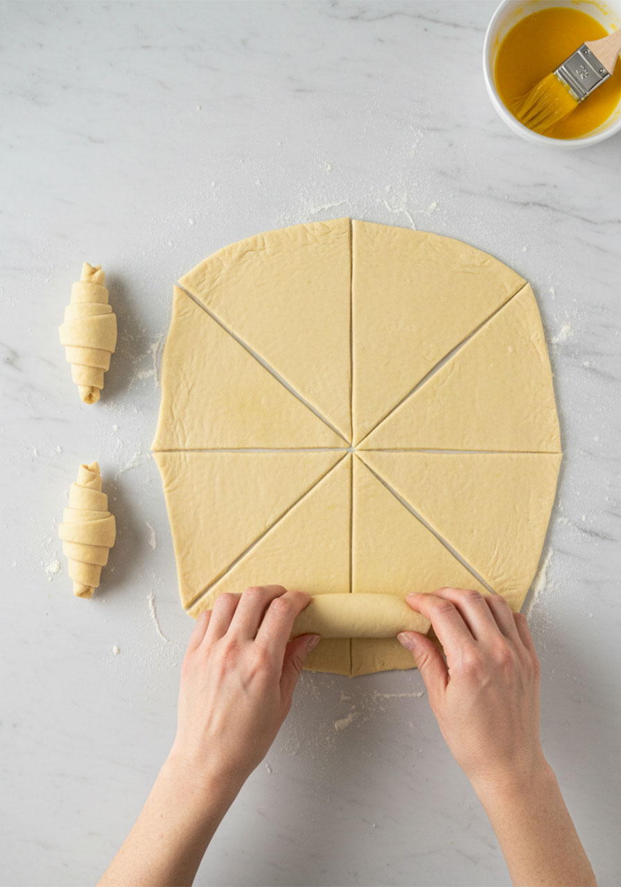 Hands rolling puff pastry triangles into mini croissant shapes on a marble countertop for easy party snacks