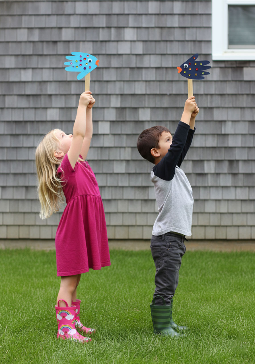 Two young children playing outdoors with their finished handprint fish puppets on popsicle sticks, smiling and holding them up against a gray shingle house