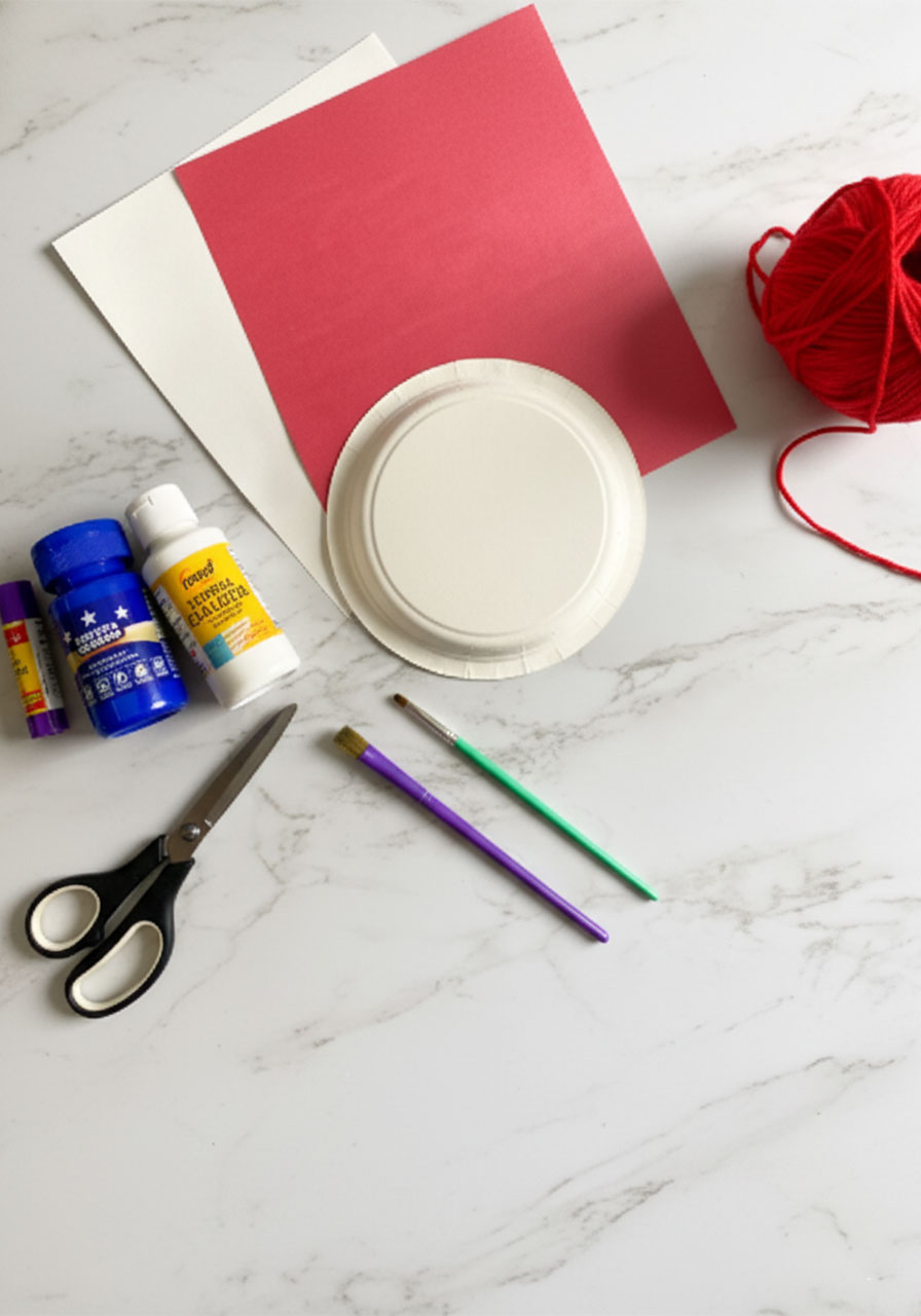 Overhead flat lay of all supplies needed for patriotic paper plate hat craft including paper plate, red and white construction paper, blue and white paint, scissors, glue stick, paintbrushes, and red yarn