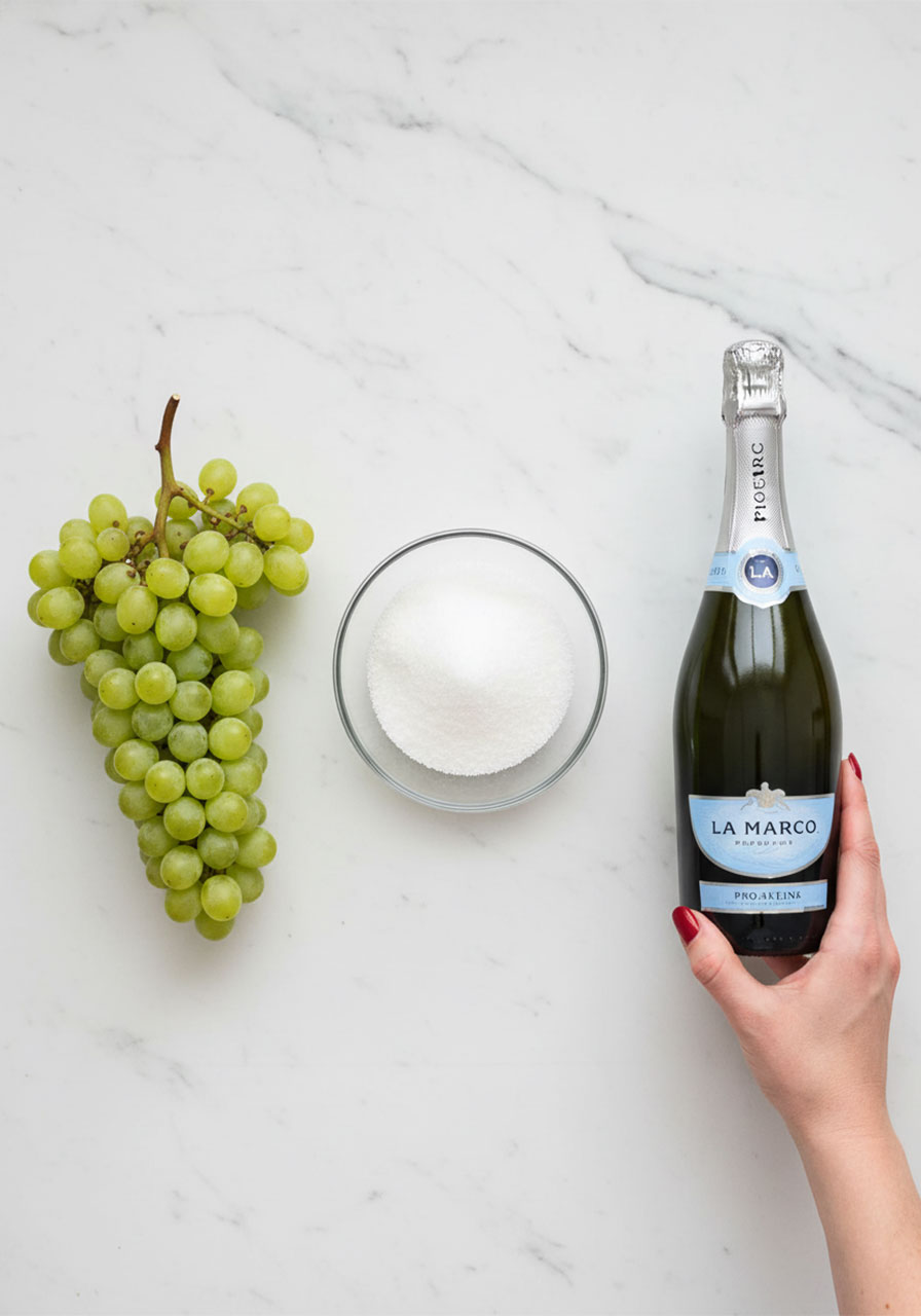 Three ingredients for prosecco grapes: fresh green grapes, cane sugar, and a bottle of prosecco on marble