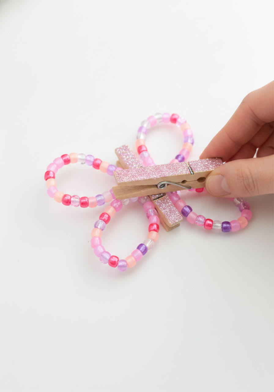Two adult hands holding a glitter clothespin open while sliding two beaded figure-eight pipe cleaner wing pieces inside to assemble a butterfly craft