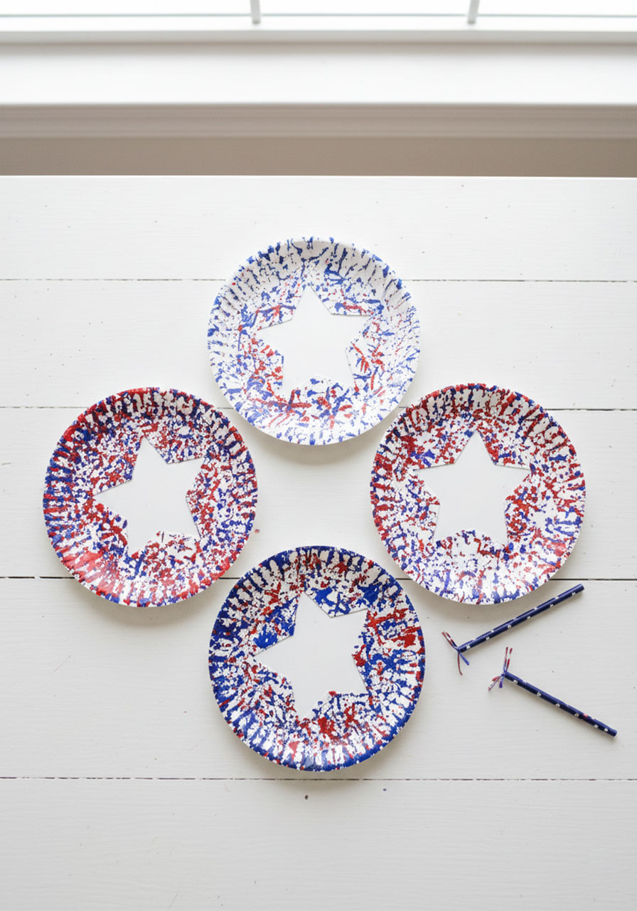 Three completed straw stamped paper plate 4th of July crafts with white star cutouts and red white and blue splatter paint on white wood table