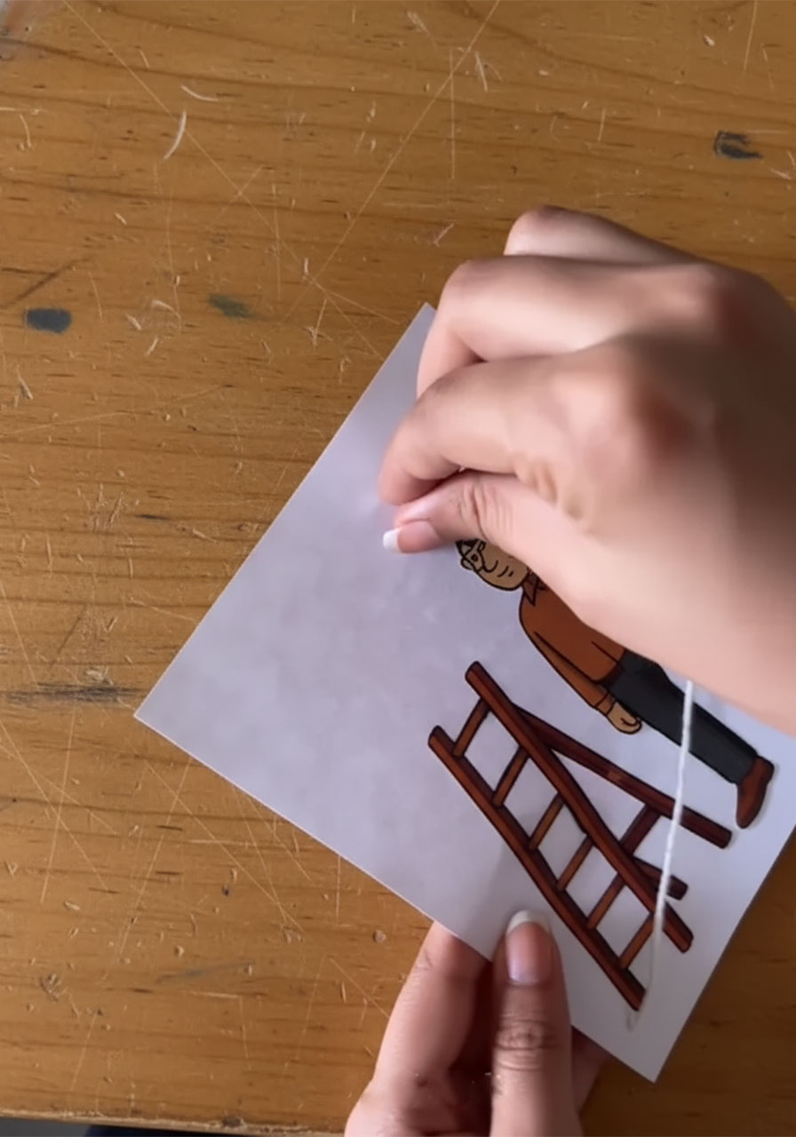 Adult hands threading white twine through two holes in cardstock to create the pull-string mechanism for a DIY Father's Day card step4a