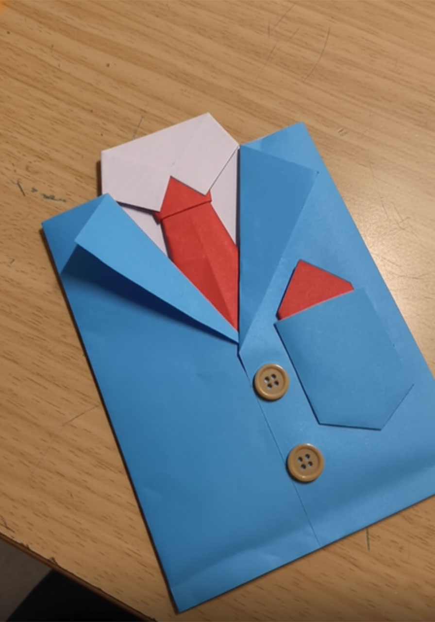 Hands gluing small wooden buttons and a red paper pocket square onto a blue paper suit jacket Father's Day card on a white table step4i
