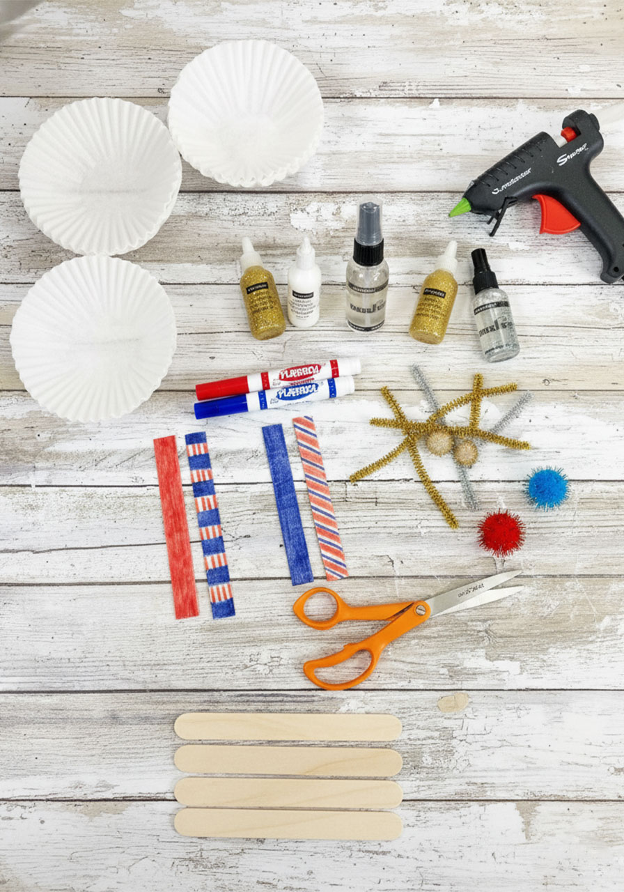 All supplies needed for coffee filter watercolor fireworks craft laid out flat — coffee filters, red and blue washable markers, spray bottle, glitter glue, craft sticks, pipe cleaners, pom poms, hot glue gun, and scissors