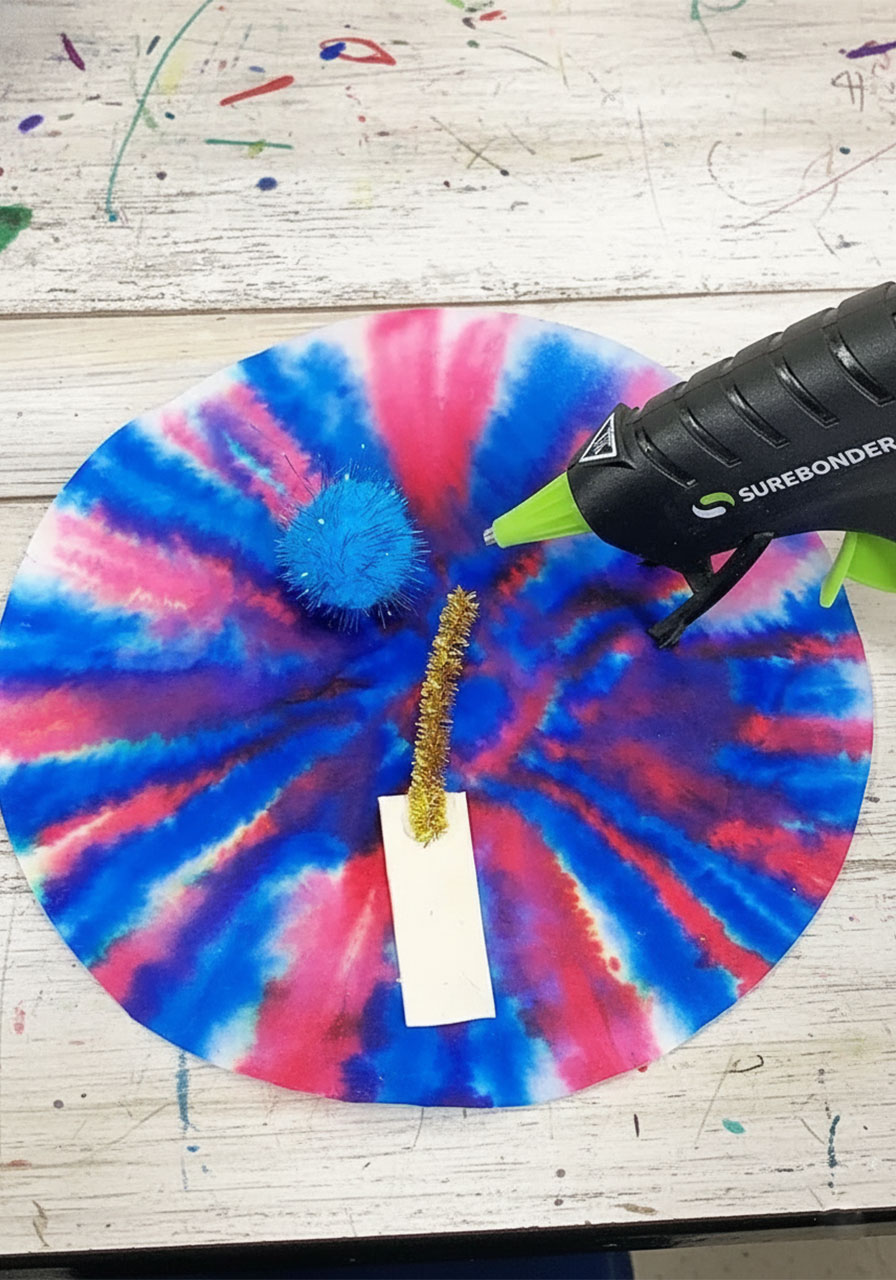 Adult using hot glue gun to attach painted craft stick and gold pipe cleaner to a dried watercolor coffee filter fireworks craft — Step 5 assembly