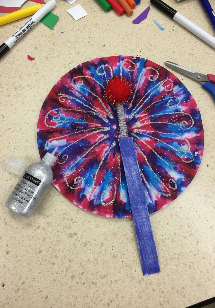 Adding gold glitter glue swirl trails radiating from the center of a coffee filter watercolor fireworks craft — Step 6 final decoration variation 1
